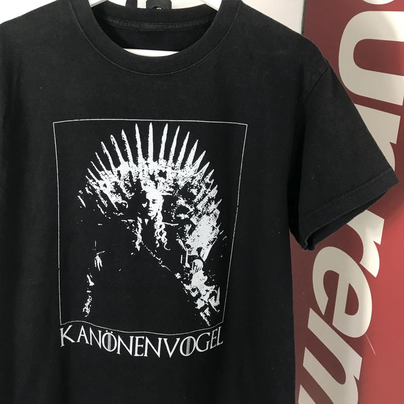 Może przedstawiać: Czarny t-shirt z białym nadrukiem przedstawiającym tron i twarz kobiety oraz napisem "KANONENVOGEL" poniżej. Koszulka ma okrągły dekolt i kr&oacute;tkie rękawy. Projekt jest wyśrodkowany z przodu.