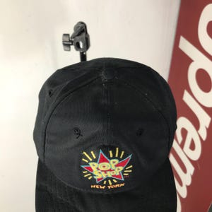 May include: Black baseball cap with a colorful "Pop Shop New York" logo embroidered on the front. The logo features a red and blue burst design with yellow accents. The cap is made of a durable fabric.