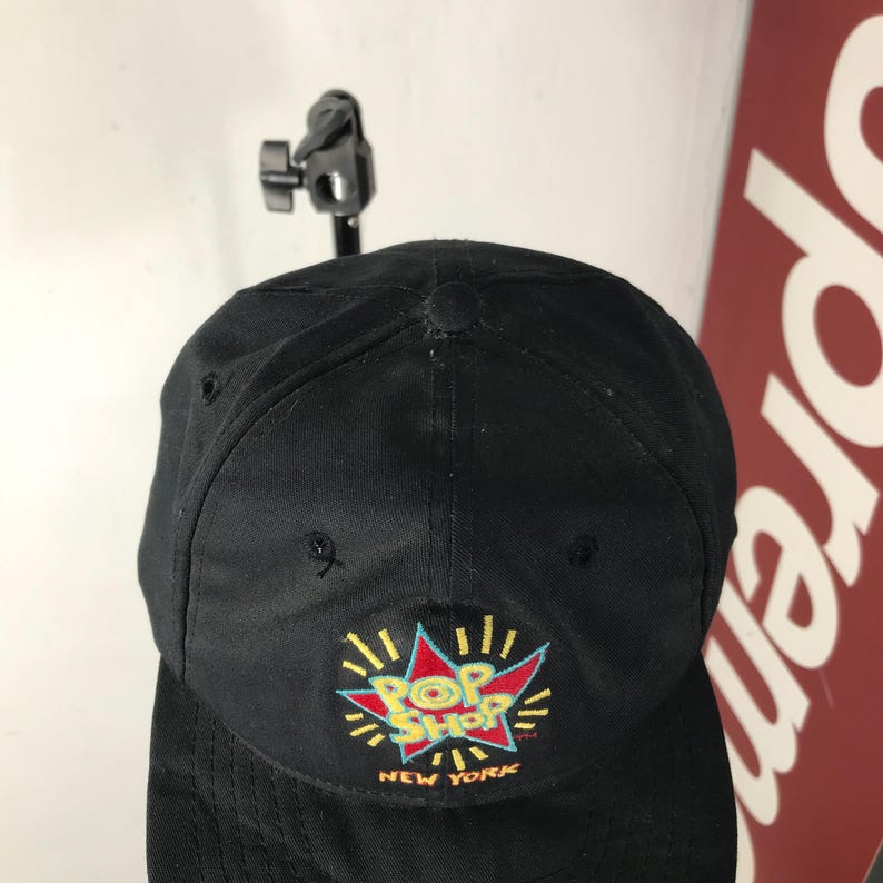 May include: Black baseball cap with a colorful "Pop Shop New York" logo embroidered on the front. The logo features a red and blue burst design with yellow accents. The cap is made of a durable fabric.
