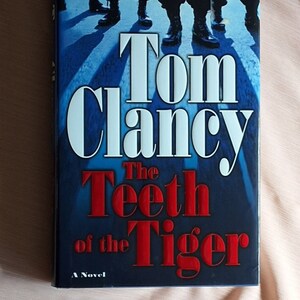 May include: A blue paperback book with the title "The Teeth of the Tiger" by Tom Clancy. The book cover features a group of people walking in a crosswalk.
