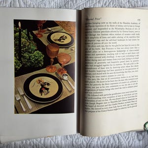 May include: A dining table setting with two plates featuring a black border and a central illustration. The table is set with silverware, amber-colored glasses, and a tall black candle. Green foliage adds a decorative touch.