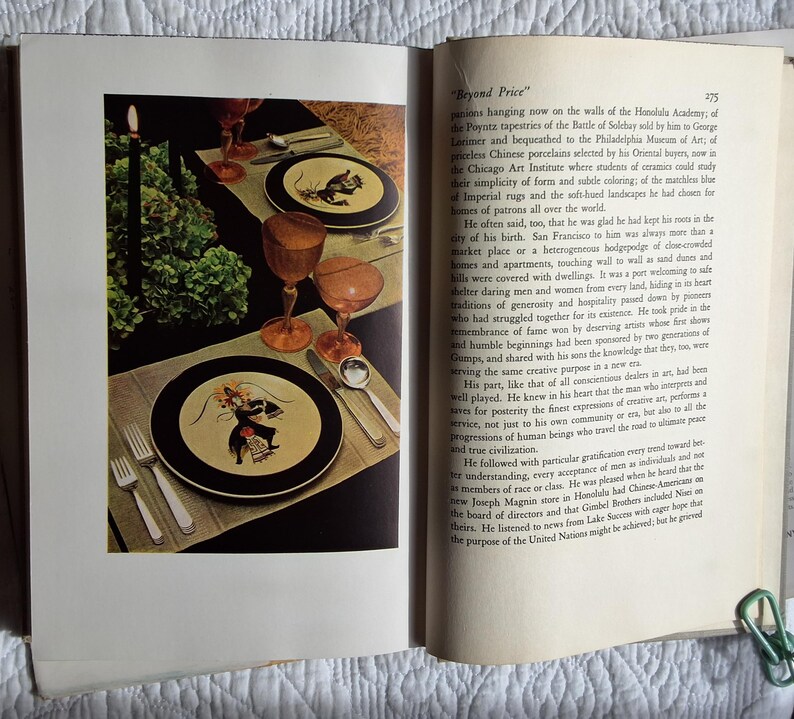 May include: A dining table setting with two plates featuring a black border and a central illustration. The table is set with silverware, amber-colored glasses, and a tall black candle. Green foliage adds a decorative touch.