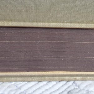 May include: A vintage book with a light tan cover and aged, purple-toned pages. The book's spine is visible, showing the binding and the edge of the pages. The cover appears to be made of a textured fabric.
