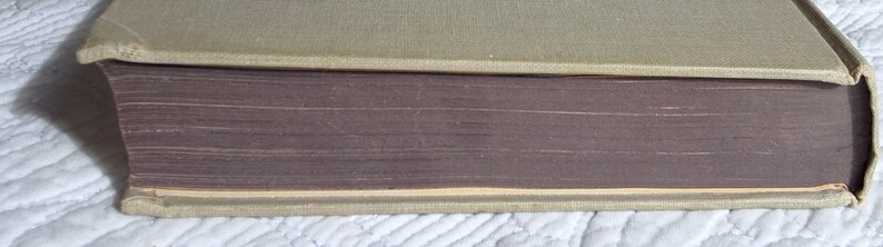 May include: A vintage book with a light tan cover and aged, purple-toned pages. The book's spine is visible, showing the binding and the edge of the pages. The cover appears to be made of a textured fabric.