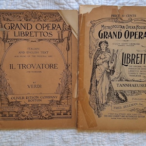 May include: Two vintage opera libretto covers. The left cover is brown with black text and illustrations. The text reads "Grand Opera Librettos, Italian and English Text and Music of the Principal Airs, Il Trovatore (The Troubadour) by Verdi, Oliver Ditson Company Boston". The right cover is beige with black text and illustrations. The text reads "Metropolitan Opera House Grand Opera, Giulio Gatti-Casazza General Manager, Libretto, The Original Italian, French or German Libretto with a Correct English Translation, Tannhaeuser, Published by Fred Rullman, Inc., 17 East 42nd Street, New York City, The Only Correct and Authorized Edition, Knabe Piano Used Exclusively".