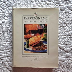 May include: A white book with a red and white logo on the front cover. The title of the book is "D'Artagnan's Glorious Game Cookbook". The book is about cooking with game meat. The cover features a photo of a sandwich with a glass of red wine.