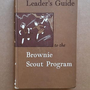 May include: A vintage brown "Leader's Guide to the Brownie Scout Program" book. The cover features illustrations of girls and text in white. The book has a worn appearance, suggesting it is an older edition.