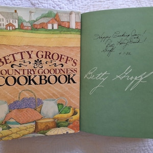 May include: A green hardcover cookbook titled "Betty Groff's Country Goodness Cookbook" with a rural scene on the front cover and a handwritten inscription on the inside cover. The inscription reads "Happy Cooking Jean!  Lots of Love, Barb!  Betty Groff 4-1-72".