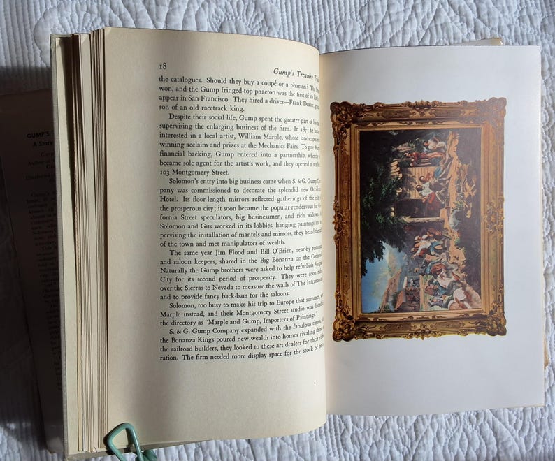May include: An open book with text on the left page and a framed painting on the right. The painting has a gold frame and depicts a scene with figures and architectural elements. A green paper clip holds the pages together.