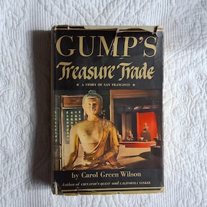 May include: Vintage book titled "Gump's Treasure Trade: A Story of San Francisco" by Carol Green Wilson. The cover features a gold-toned Buddha statue and a red-lit display case. The book has a black background with gold lettering.