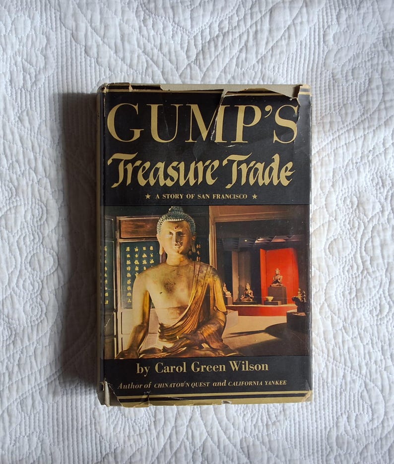 May include: Vintage book titled "Gump's Treasure Trade: A Story of San Francisco" by Carol Green Wilson. The cover features a gold-toned Buddha statue and a red-lit display case. The book has a black background with gold lettering.