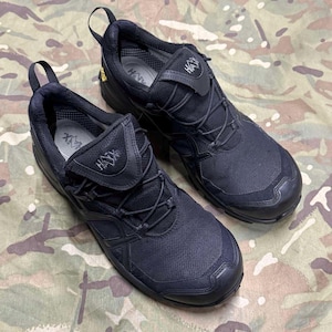 Könnte beinhalten: Ein Paar schwarze, flache Sportschuhe mit einem Obermaterial aus Mesh und Synthetik, einem Schnellschnürsystem und einem gepolsterten Schaft. Die Zunge zeigt das HAX-Logo und ein kleines gelbes Etikett ist an der Fersenseite sichtbar.