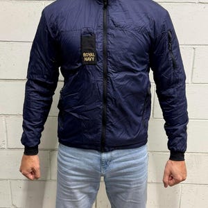 Puede incluir: Una chaqueta azul marino ligera con cuello alto y cremallera frontal negra completa. La chaqueta presenta un parche rectangular negro en el pecho derecho con el texto ROYAL NAVY en dorado y bolsillos negros con cremallera en las mangas.