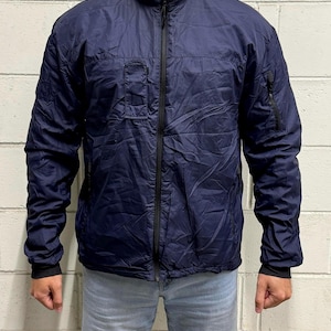 Pode incluir: Um casaco leve azul-marinho com gola alta, fecho de correr frontal preto a todo o comprimento e mangas compridas com punhos canelados pretos. O casaco inclui um bolso vertical com fecho no peito esquerdo e na manga esquerda.
