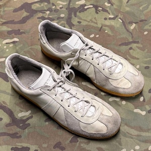 May include: A pair of low-top sneakers featuring a white leather upper with light gray suede overlays on the toe and side panels. The shoes include white laces, a padded gray interior lining, and a flat gum rubber sole for a classic athletic look.