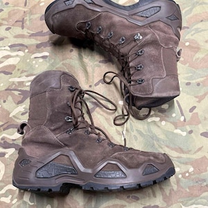 Könnte beinhalten: Ein Paar taktische Stiefel aus braunem Wildleder mit hohem Knöchelschutz, dunkelbraunen Gummisohlen mit geometrischen Texturdetails, Metallösen für die Schnürung und einer verstärkten Fersenstruktur für den robusten Außeneinsatz.