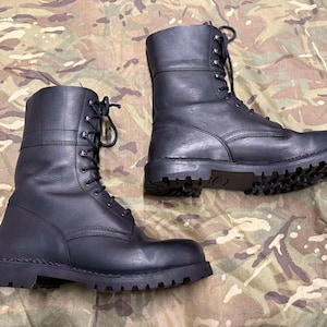 Könnte beinhalten: Ein Paar schwarze Leder-Kampfstiefel mit Schnürung. Die Stiefel haben einen hohen Schaft und robuste Sohlen. Die Stiefel werden auf einer Oberfläche mit Tarnmuster präsentiert.