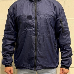 Pode incluir: Um casaco leve azul-marinho com fecho de correr preto a todo o comprimento, gola subida e mangas compridas com punhos pretos. O casaco inclui um bolso vertical com fecho na manga esquerda e um bolso retangular com aba no peito.