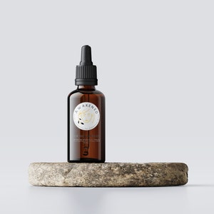 May include: A brown glass bottle with a black dropper top and a white label with the text "AWAKENED CO. Nourishing Skin Essentials" and a gold illustration of a woman's face. The bottle is sitting on a round, grey stone.