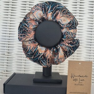May include: A black and teal hair scrunchie with a peacock feather pattern. The scrunchie is on a black stand and has a brown tag that says "Handmade with Love NATSKI www.handmadebynatski.com".