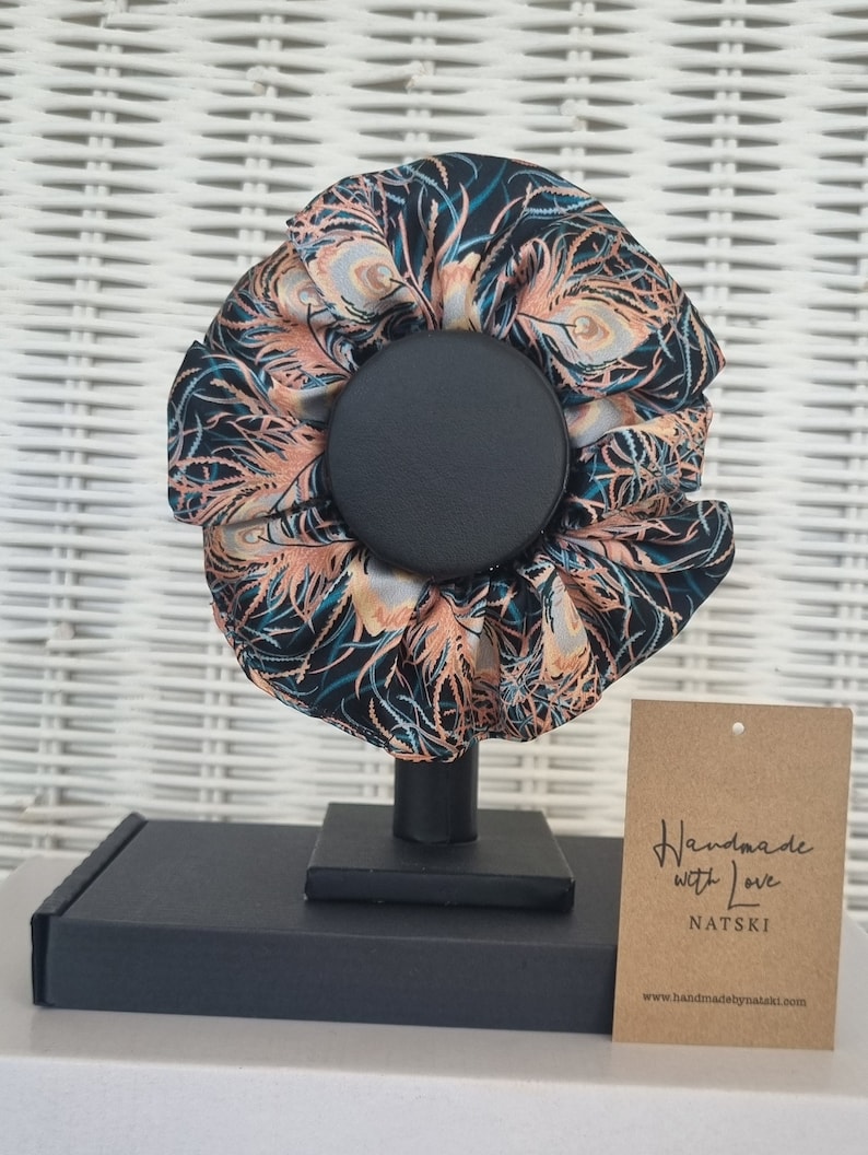 May include: A black and teal hair scrunchie with a peacock feather pattern. The scrunchie is on a black stand and has a brown tag that says "Handmade with Love NATSKI www.handmadebynatski.com".