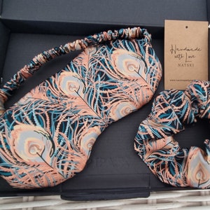 May include: A black box with a peach and teal peacock feather print sleep mask and hair scrunchie. The box has a tag that reads "Handmade with Love NATSKI" and a website address.