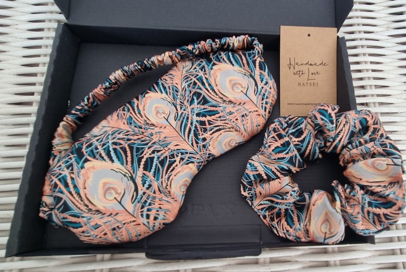May include: A black box with a peach and teal peacock feather print sleep mask and hair scrunchie. The box has a tag that reads "Handmade with Love NATSKI" and a website address.