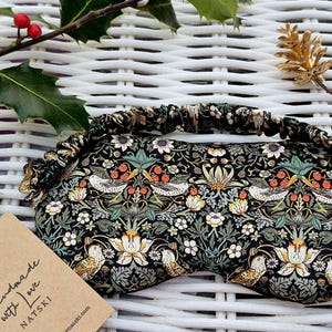May include: A black eye mask with a floral and bird design. The mask features a patterned elasticated band and is decorated with white flowers, green leaves, and red berries. A tag reads "Handmade with Love".