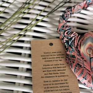 May include: A brown paper tag with text that reads "Unwind, relax your mind and let the lavender help to soothe away the stresses of the day. Pillows are made from Luxurious Silk Satin and are filled with Lavender and flax Seeds. Great for relaxation & meditation. Heat on radiator for few mins or in microwave for 15-20 seconds, ensuring pillow isn't too hot before placing over eyes. Take some deep breaths and enjoy!" The tag is attached to a pink and blue patterned eye pillow.