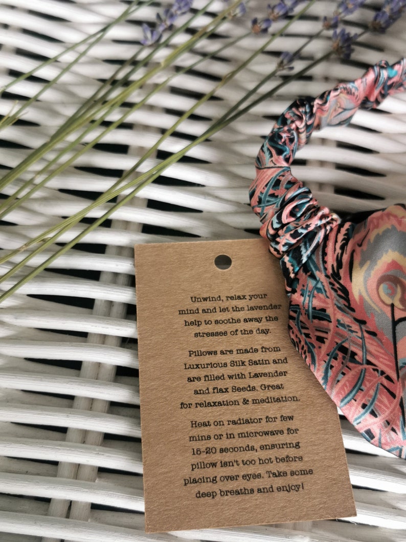 May include: A brown paper tag with text that reads "Unwind, relax your mind and let the lavender help to soothe away the stresses of the day. Pillows are made from Luxurious Silk Satin and are filled with Lavender and flax Seeds. Great for relaxation & meditation. Heat on radiator for few mins or in microwave for 15-20 seconds, ensuring pillow isn't too hot before placing over eyes. Take some deep breaths and enjoy!" The tag is attached to a pink and blue patterned eye pillow.