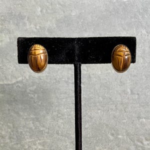 Könnte beinhalten: Ein Paar oval geformte Ohrringe aus braunem Tigerauge-Edelstein, die in Form von Skarabäus-Käfern geschnitzt sind. Die Ohrringe haben eine glatte, polierte Oberfläche mit natürlicher Chatoyance-Bänderung und sind auf einem schwarzen Samtständer.