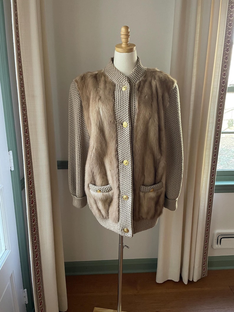 May include: A beige knitted cardigan with a fur panel down the front and gold buttons. The cardigan is on a white mannequin.