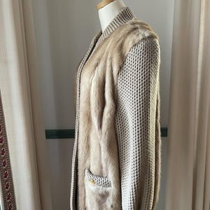 May include: A beige knitted cardigan with a fur panel down the front. The cardigan has a zipper closure and a pocket on the left side.