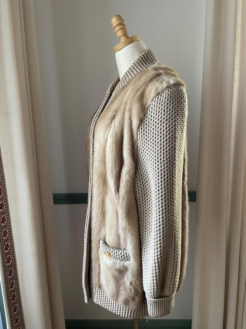 May include: A beige knitted cardigan with a fur panel down the front. The cardigan has a zipper closure and a pocket on the left side.