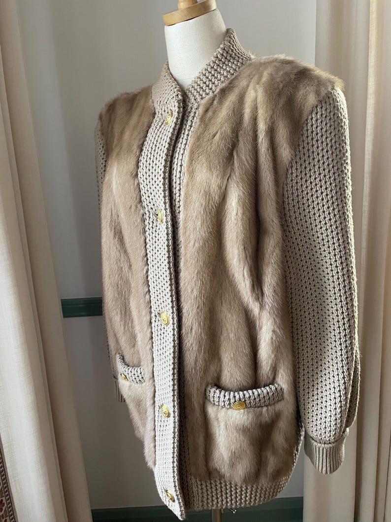 May include: A beige knitted cardigan with fur panels on the front and sleeves. The cardigan has a button closure and two pockets.