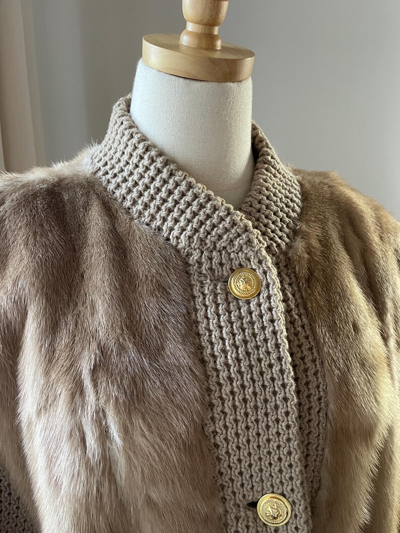 May include: A brown fur jacket with a knitted collar and gold buttons. The jacket is on a white mannequin.