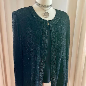 May include: A black beaded jacket with long sleeves and a collarless design. The jacket is displayed on a mannequin with a silver necklace and pendant. The jacket's surface is covered in small, dark beads, creating a textured appearance.