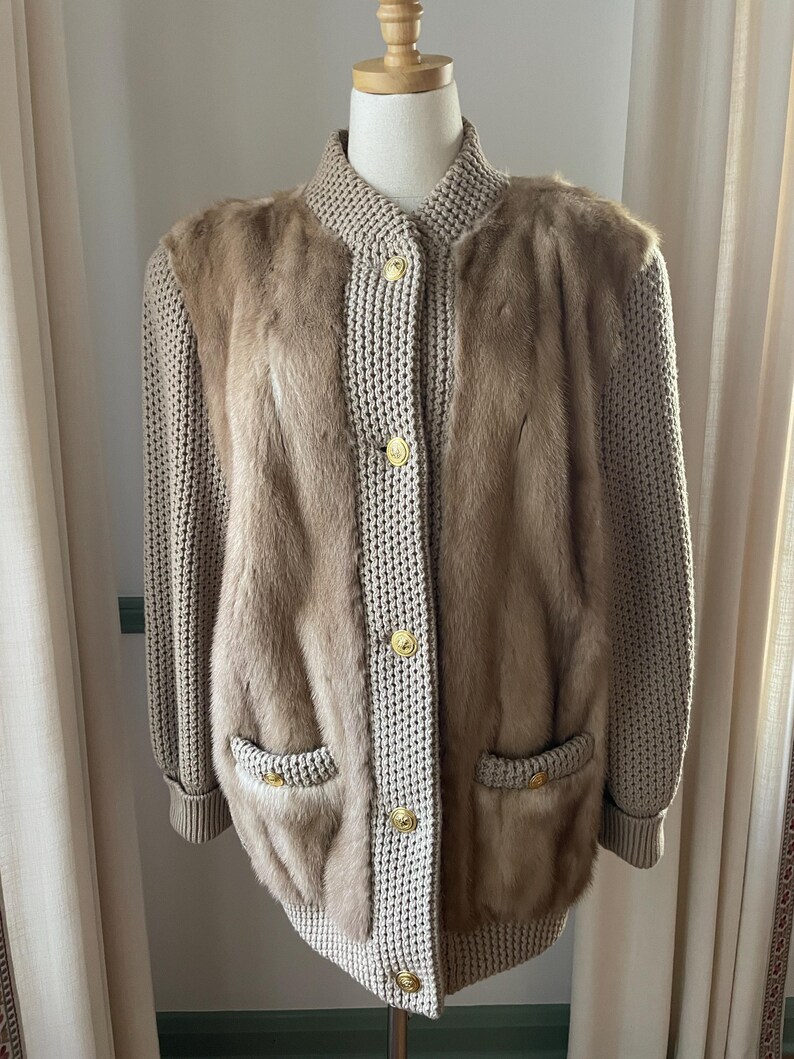 May include: A beige knitted cardigan with fur detailing on the front and sleeves. The cardigan has a button closure and two pockets.