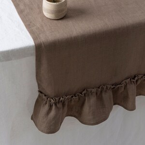 May include: A brown linen table runner with a ruffled edge. A small, beige ceramic vase holds a dried flower. The runner is draped over a white tablecloth, creating a neutral, rustic aesthetic.