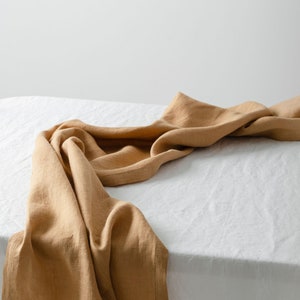 May include: A draped, golden-brown linen table runner. The fabric is soft and textured, with folds and shadows that add depth. The runner is placed on a white surface, creating a contrast in color and texture. Ideal for home decor.