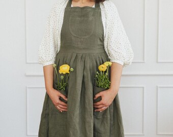 Op de afbeelding: Een vrouw draagt een groene linnen schort met twee zakken, elk met een klein boeket gele bloemen. Het schort heeft een geplooide rok en een grote zak aan de voorkant.