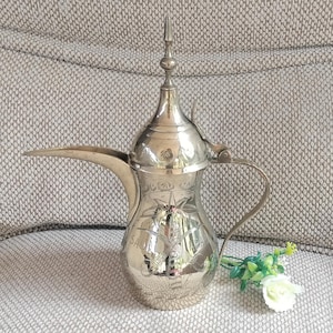 May include: Silver-colored, ornate coffee pot with a long spout and a handle. The pot has engraved designs and a pointed lid. The word "SAUDI" is visible on the pot. A small bouquet of white and green flowers rests near the pot.