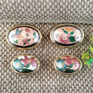 May include: Four oval-shaped trinket boxes with gold-colored rims. The boxes feature floral and bird designs in pink, green, and blue enamel. The boxes are arranged on a textured, tan surface.