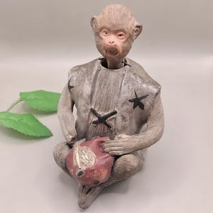 May include: A ceramic monkey figurine in a seated position, holding a red fruit. The monkey wears a gray vest with black star and cross accents. The figurine has a light brown face and hands.