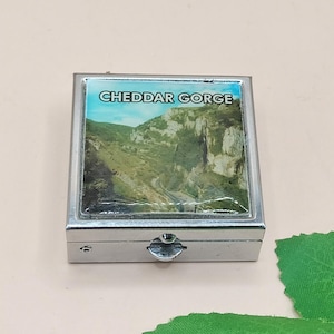 May include: A small, square, silver-colored pill box with a hinged lid. The lid features a scenic image of Cheddar Gorge, with green hills and a winding road. The text "CHEDDAR GORGE" is printed at the top of the image.