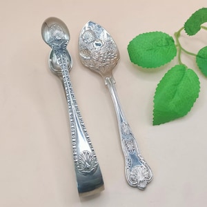 May include: Two antique silver serving spoons. One spoon is a sugar tong with a shell design on the handle and the spoon end. The other spoon has an ornate floral design on the spoon end and handle. The spoons are on a light pink surface.