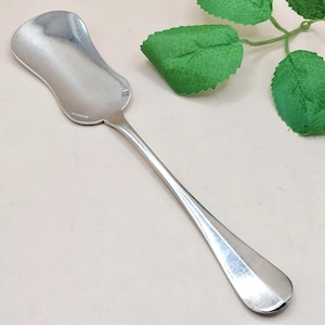 Könnte beinhalten: Ein silberfarbener Zuckerlöffel mit einer breiten, gebogenen Schale und einem langen, schlanken Stiel. Der Löffel hat ein rechteckiges Detail auf der Schale. Der Löffel ist wahrscheinlich aus Edelstahl oder einem ähnlichen Metall gefertigt. Der Löffel ist zum Servieren von Zucker oder anderen körnigen Artikeln bestimmt.