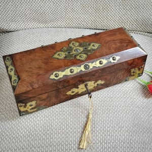 May include: A rectangular wooden box with a polished burl wood finish, featuring ornate brass decorative plates on the lid and front. A brass key is inserted into the front lock, attached to a long, thin, gold-colored decorative tassel.