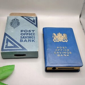 May include: Two vintage Post Office Savings Bank boxes. One is a light blue cardboard box with the text "POST OFFICE SAVINGS BANK". The other is a blue leather-bound book with gold lettering and a royal crest.