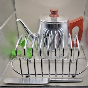 May include: A vintage tea set featuring a silver teapot, a chrome toast rack, and a butter knife. The teapot has a wooden handle and lid knob. The toast rack holds several slices of toast. The butter knife is silver.